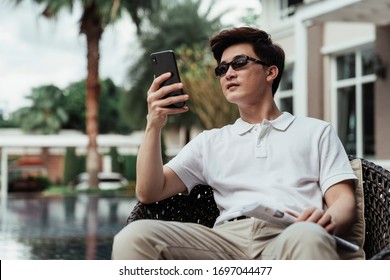 Young Asian Guy On White Shirt Wearing Sunglasses Using Mobile Phone At Resort Villa.