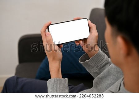 A young asian guy using his smartphone, watching video or playing mobile game. mobile phone white screen mockup. close-up, back view