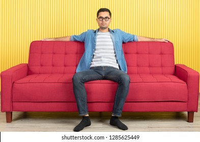 Young Asian handsome bearded man, wearing eyeglasses in denim shirt, sitting on center of red sofa, arms wide spread in confident, in modern living room, bright yellow stripe wall background