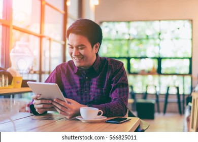 Young Asian Handsome Businessman Smiling While Reading His Tablet With A Cup Of Coffee And Smartphone In Coffee Cafe. Portrait Of Asian Business Man Reading Message With Tablet And Smart Phone.