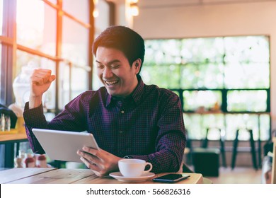 Young Asian Handsome Businessman Smiling Cheering While Reading His Tablet With A Cup Of Coffee And Smartphone In Coffee Cafe. Portrait Of Asian Business Man Reading Message With Tablet.