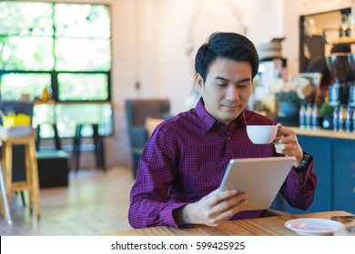 Young Asian Handsome Businessman Smiling While Reading His Tablet With A Cup Of Coffee And Smartphone In Coffee Cafe. Portrait Of Asian Business Man Reading Message With Tablet And Smart Phone In Shop