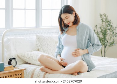 Young Asian happy pregnant woman is sitting and relaxing on bed and and touching her belly. Family plan, pregnancy, motherhood, people and expectation concept - Powered by Shutterstock