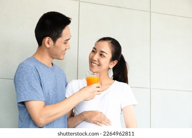 Young Asian healthy family, husband preparing orange juice for lovely pregnant woman smile and drink orange juice while standing in modern living room. New couple health care concept. - Powered by Shutterstock