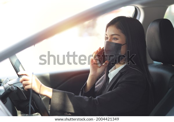 Young asian healthy\
woman in business black suit with protect mask for healthcare in\
automobile and using smartphone and driving car. New normal and\
social distancing concept