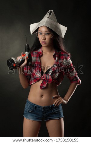 Young asian high fashion glamour model in  shorts, safe glasses and newspaper hat holding electric drill in her hand over dark background