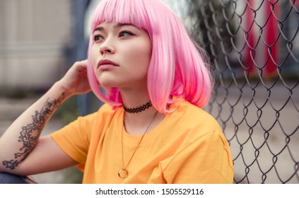 Young Asian Hipster With Dyed Pink Hair Looking Away And Thinking While Sitting Near Fence On Modern City Street