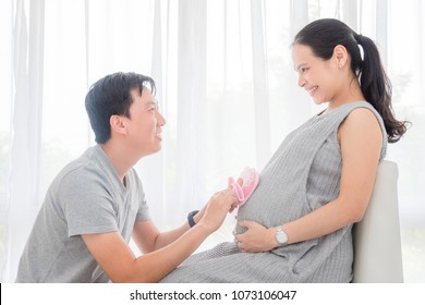 Young asian husband put small baby shoes on his pregnant wife's belly and smiles - Powered by Shutterstock