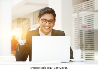 Young Asian Indian business people celebrating success, using a laptop at cafe, with a cup of coffee. India male business man, modern building with beautiful golden sunlight as background.