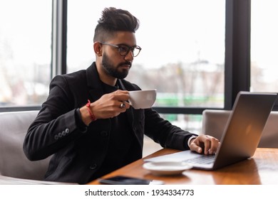 Young Asian Indian businessman using a notebook computer or laptop during office break at cafe, relaxing with a cup of coffee. India male business man, real modern office building as background. - Shutterstock ID 1934334773