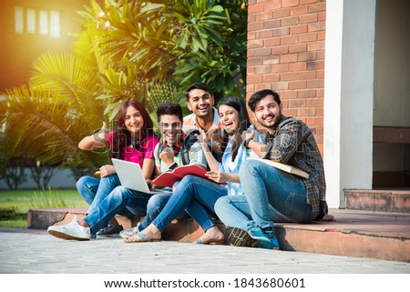 Young Asian Indian college students reading books, studying on laptop, preparing for exam or working on group project while sitting on grass, staircase or steps of college campus