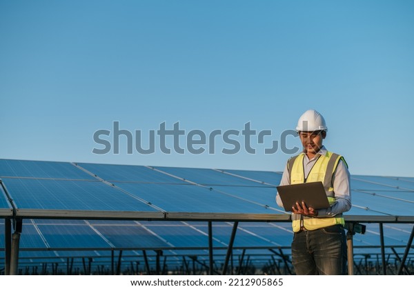 Young Asian Inspector Engineer man use laptop\
computer working at solar farm, Technician, supervisor male in\
white helmet Checking operation of sun and photovoltaic solar panel\
in station, copy space