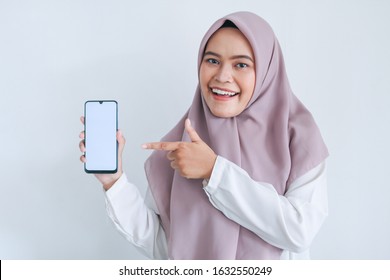Young Asian Islam woman wearing headscarf is pointing finger on white screen of phone with smile and happy feeling. Indonesian woman on gray background.