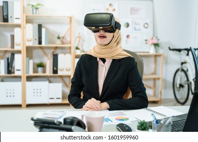 Young Asian Islam Woman Worker In Professional Black Suit Having Fun In Virtual Reality Goggle In Office. Female Muslim Lady Sitting At Work Desk In Modern Studio And Laughing With Vr Headset