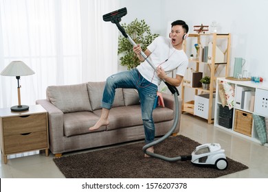 Young Asian Japanese Man Having Fun During Cleaning House. Handsome Guy Relax Playing With Vacuum Cleaner At Living Room While Wearing Headphones And Dancing With Music. Male Household Moving Body