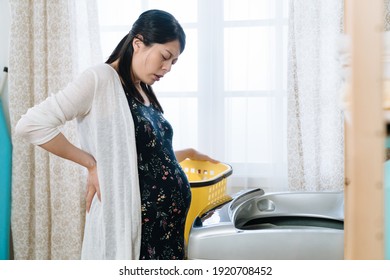 Young Asian Japanese Maternity Lady Suffering From Back Pain While Doing Laundry By Washing Machine. Beautiful Pregnant Woman Housewife With Hurting Waist And Big Abdomen Frowning Face Do Housework