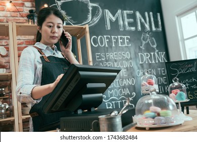 young asian japanese woman staff working in cafe store. lady waitress talking on cellphone while customer taking order. female barista using point of sale terminal and mobile phone in coffee shop.
