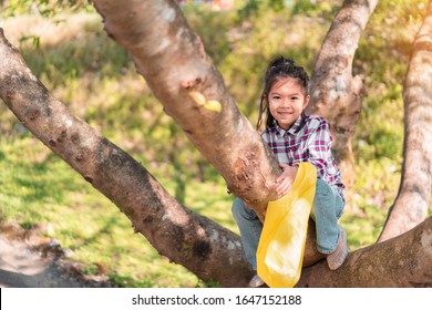 Young Asian Kid Girl Playing Climbing To A Tree In A Park Outdoor, Selective Focus.