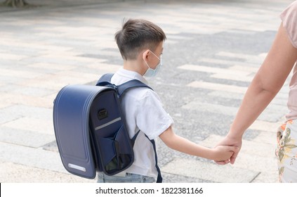 The Young Asian Kid Is Walking To School With The Parent And Also Carrying The Big And Heavy School Backpack. Young Student With The Large Bag On The Shoulder Holding Hand With His Mom.