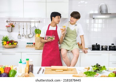 Young Asian LBGT Couple Toast Wine And Hold Salad To Eat After Cooking At Kitchen. Lovely Gay Couple. Healthy Breakfast Eating Lifestyle For Same Sex Family In Morning.