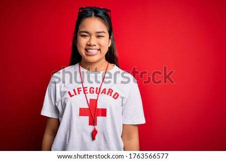 Young asian lifeguard girl wearing t-shirt with red cross using whistle over isolated background with a happy and cool smile on face. Lucky person.