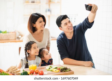 Young Asian Love Family Are Preparing The Breakfast In The Kitchen. Father Take A Photo Selfie By Phone. Excited Smiling And Felling Happy. Parent Teach Daughter To Cooking Food On The Day At Home. 