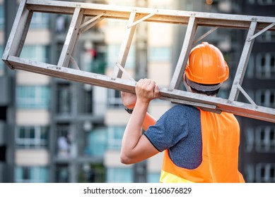 Young Asian Maintenance Worker Man With Orange Safety Helmet And Vest Carrying Aluminium Step Ladder At Construction Site. Civil Engineering, Architecture Builder And Building Service Concepts