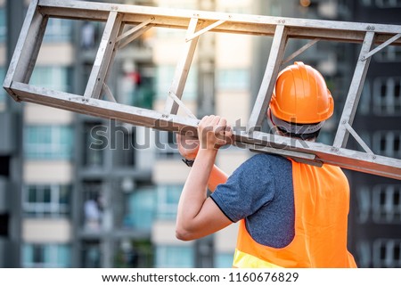 Young Asian maintenance worker man with orange safety helmet and vest carrying aluminium step ladder at construction site. Civil engineering, Architecture builder and building service concepts