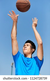 Young Asian Male Basketball Player Making A Jump Shot Against Blue Sky Background.