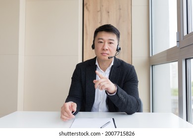 Young Asian Male Businessman Talking Online On Video Call, Conference Or Meeting Looking At Camera. Webcam View. Asia Man In Business Suit Indoors. Office Negotiations Distance Consultation Or Advice