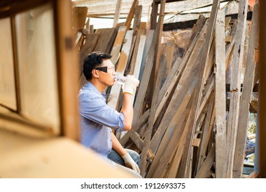 Young Asian male carpenter wearing safety goggles and gloves sitting drinking water from bottle while taking a break after woodworking in wood factory with many vertically stacked wood in background.
