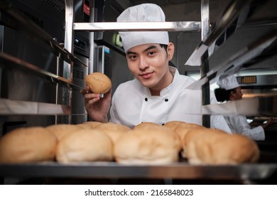Joven chef asiático con uniforme blanco para cocinar y sombrero que muestra una bandeja de pan fresco y sabroso con una sonrisa, mirando a la cámara, feliz con sus productos de comida horneada, trabajo profesional en la cocina de acero inoxidable.