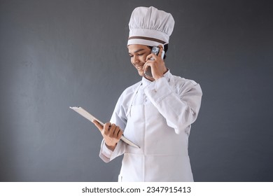 Young Asian male chef in white apron and toque holding a clipboard while having a phone call isolated over grey background - Powered by Shutterstock