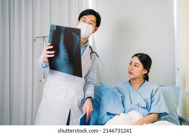 Young Asian male doctor with face mask and stethoscope in labcoat explaining and reviewing x-ray report to patient sitting in hospital bed - Powered by Shutterstock