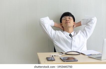 Young Asian Male Doctor Sitting At Desk Hands Under His Head Relaxing With Eyes Close.  Tired Handsome Nurse In A White Coat Takes A Break Sits Relax. Isolated On White Background Copy Space.