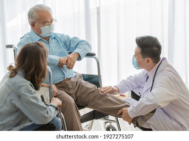 A Young Asian Male Doctor Wearing White Lab Coat And Face Mask Checking Left Knee Of Old Fat Gray Hair Patient Wearing Light Blue Shirt And Brown Pants While He Sitting On Wheelchair When His Daughter