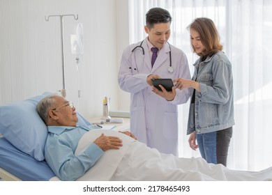 A Young Asian Male Doctor Wearing White Lab Coat And Stethoscope Using Black Tablet Explaining Patient Test Result To Female Relative When Old Fat Patient Wearing Eyeglasses And Light Blue Shirt 