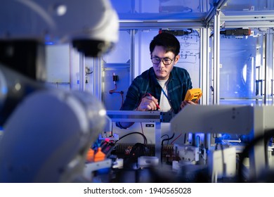 Young Asian male electrical engineer in glasses using a digital multimeter in hand checking voltage to fix an industrial machine with a blurred of automation robotic arm machine in the foreground.