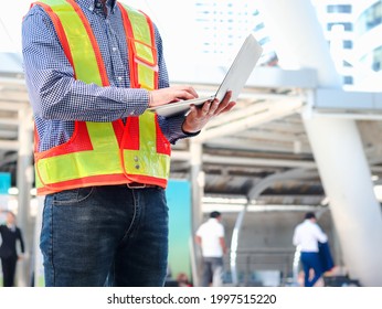 Young Asian Male Engineer Wearing Safety Vest And Helmet, Standing And Using Laptop Computer At Modern City, Young Staff Man Worker Working At The Street Outside.