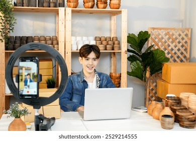 Young Asian male entrepreneur using a laptop and smartphone to live stream from his home studio, showcasing handcrafted pottery products with a selfie light setup - Powered by Shutterstock