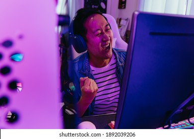 Young Asian Male Gamer Won And Victory The Winning Game With Headset And Pc Computer. Overjoyed Live Streamer Streaming Video Game In Low Light Room. Excited Gaming Influencer Smiling. With Copy Space