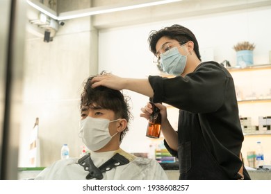 Young Asian male hairdresser in glasses spraying water on hair of Asian male client sitting on hairdressing chair before haircut in barber shop. Both of them wearing face masks for protective Covid-19 - Powered by Shutterstock