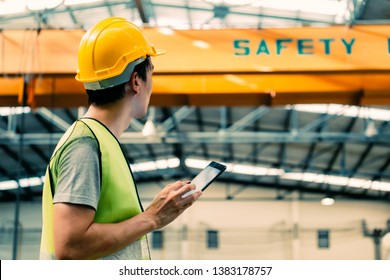 Young Asian Male Heavy Industral Worker Using A Digital Tablet Inside Manufacturing And Factory Site With Crane Behind