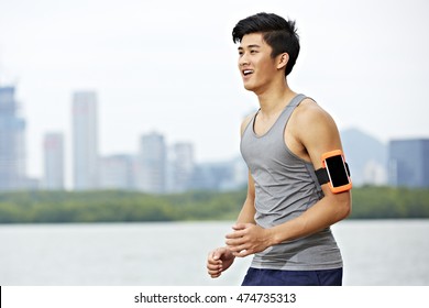 Young Asian Male Jogger With Fitness Tracker Attached To Arm Running With Skyline In The Background.