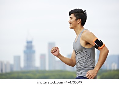 Young Asian Male Jogger With Fitness Tracker Attached To Arm Running With Skyline In The Background.