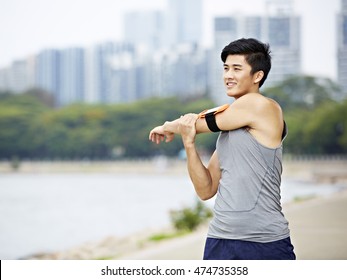 Young Asian Male Jogger With Fitness Tracker Attached To Arm Warming Up By Stretching Arms And Upper Body Before Running, City Skyline In The Background.