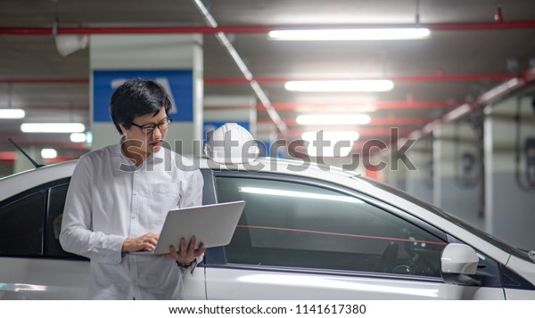 Young Asian male\
mechanical engineer working with laptop computer while standing by\
the car and white safety helmet. Mechanical engineering or building\
construction concepts