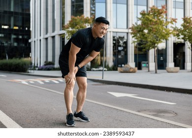 Young Asian male runner standing in the middle of the street bent over, holding his leg and knee, pulled a muscle, hiding and suffering from pain. - Powered by Shutterstock