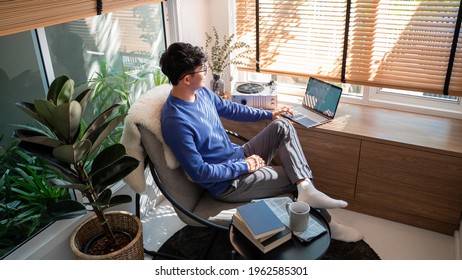 Young asian male tech user relaxing  holding laptop computer and looking at the screen in living room, Remote Job or work from home concept