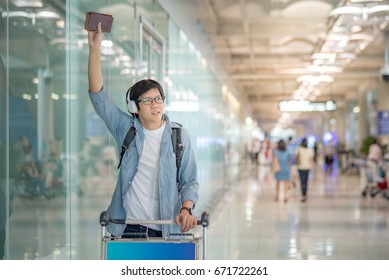 Young Asian Man With Airport Trolley Waving His Hand In The International Airport Terminal, Arrival From Travel Abroad